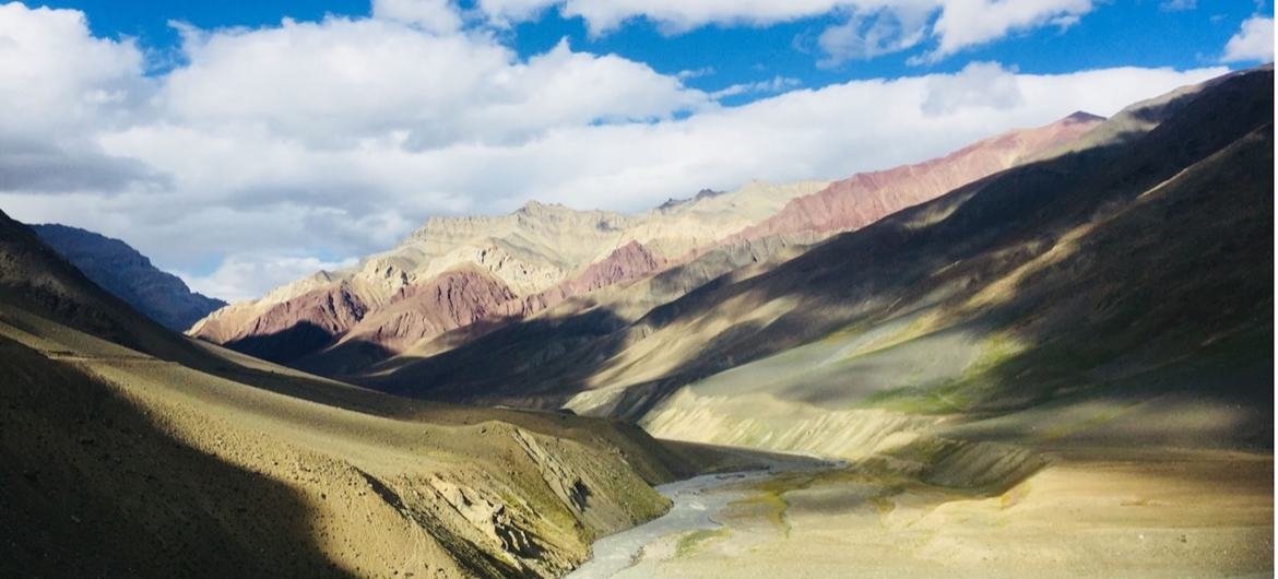 Uma vista panorâmica do vale de Spiti, a primeira reserva de biosfera do deserto frio de alta altitude da Índia, mostrando cadeias de montanhas coloridas e um rio sinuoso sob um céu azul brilhante com nuvens.