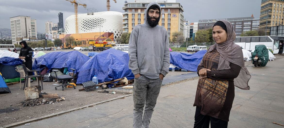 Um homem e uma mulher em frente a tendas montadas no centro da cidade, com guindastes de construção e edifícios ao fundo, representando pessoas deslocadas que vivem em condições difíceis no Líbano.